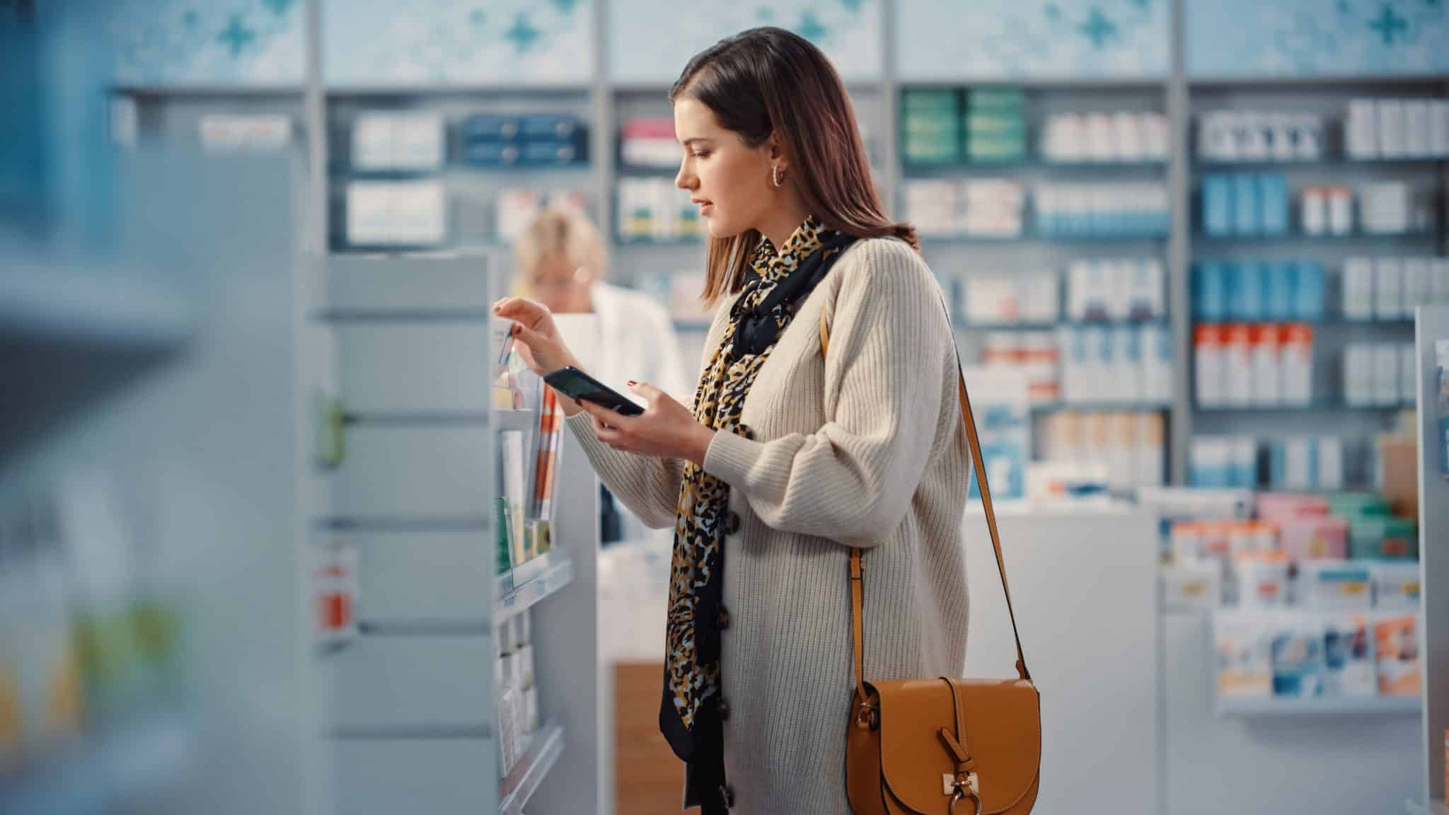 Pharmacy Drugstore: Portrait of Beautiful Young Woman Uses Smartphone, Searches to Buy Best Medicine, Drugs, Vitamins, Supplements. Shelves full of Health Care, Welness, Beauty, Cosmetics Products